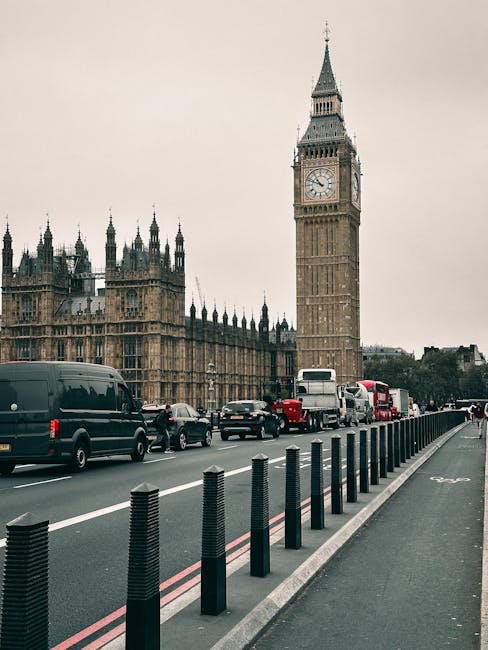 A city street scene in London featuring the iconic Big Ben clock tower and the Houses of Parliament in the background. In the foreground, a row of black bollards separates the main traffic lane from t